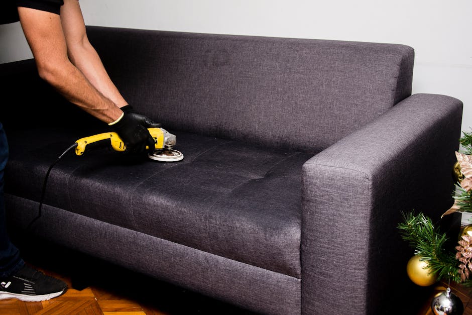 Close-up of a hand vacuuming a green upholstered armchair in a living room, with a dark gray sofa visible in the background. The room is well-lit with natural light, highlighting the clean, textured f