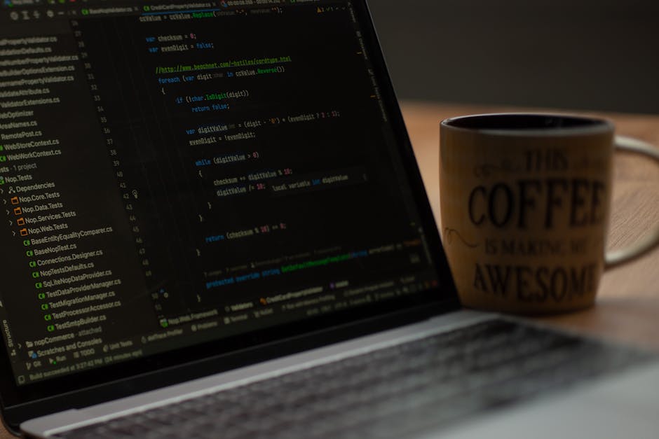 A close-up image of a laptop computer displaying lines of programming code on a dark background, with colorful syntax highlighting. Positioned to the left of the laptop is a ceramic coffee mug with a 
