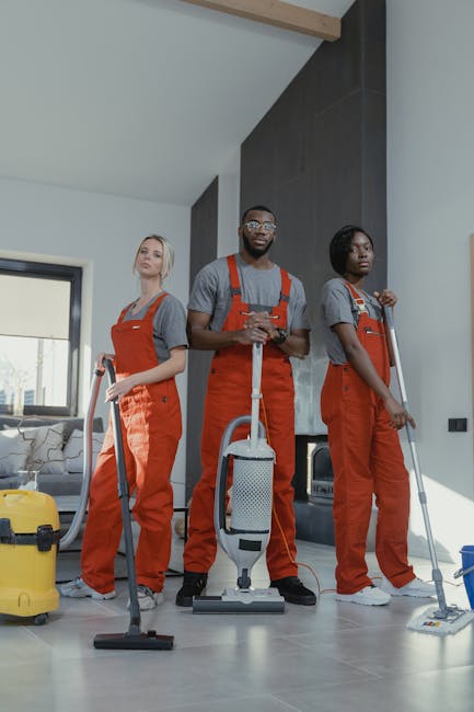 Two professional cleaning staff members from Carpet Cleaning SW4 standing indoors against a plain white wall, dressed in beige cleaning uniforms with blue badges. One is holding a vacuum cleaner while
